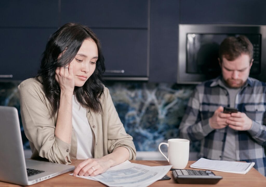 An international student sitting on a table, with hand in her jaw as she stare at a paper. She's deliberating over student loans alternative. Her laptop, coffee cup and calculator is on the table, and a man is at a far corner of the building staring at his phone.